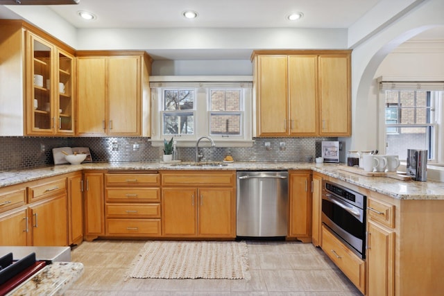 kitchen featuring glass insert cabinets, light stone counters, appliances with stainless steel finishes, arched walkways, and a sink