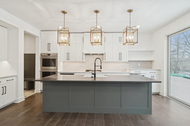 kitchen with wood finish floors, an island with sink, backsplash, appliances with stainless steel finishes, and white cabinets