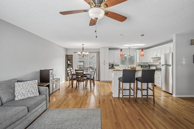 living area with baseboards, a textured ceiling, light wood-style flooring, and ceiling fan with notable chandelier