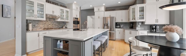 kitchen with light wood-style flooring, a center island with sink, under cabinet range hood, backsplash, and appliances with stainless steel finishes