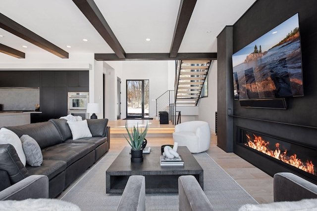 living room featuring beam ceiling, recessed lighting, stairs, light wood-style floors, and a glass covered fireplace