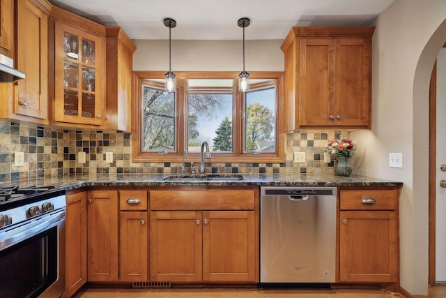 kitchen featuring dark stone countertops, arched walkways, a sink, stainless steel appliances, and brown cabinets