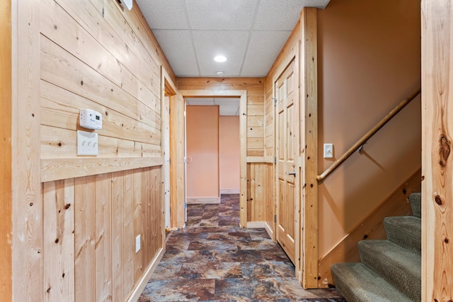 hallway with stairs, a paneled ceiling, wood walls, and stone finish flooring