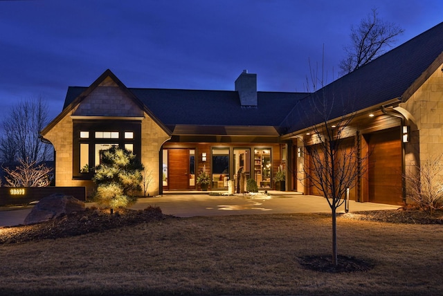 back of house at twilight with a chimney, a shingled roof, and stone siding