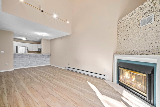 unfurnished living room with light wood-style floors, baseboard heating, a glass covered fireplace, and a towering ceiling