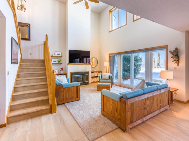 living area with a ceiling fan, a glass covered fireplace, stairway, a high ceiling, and light wood finished floors