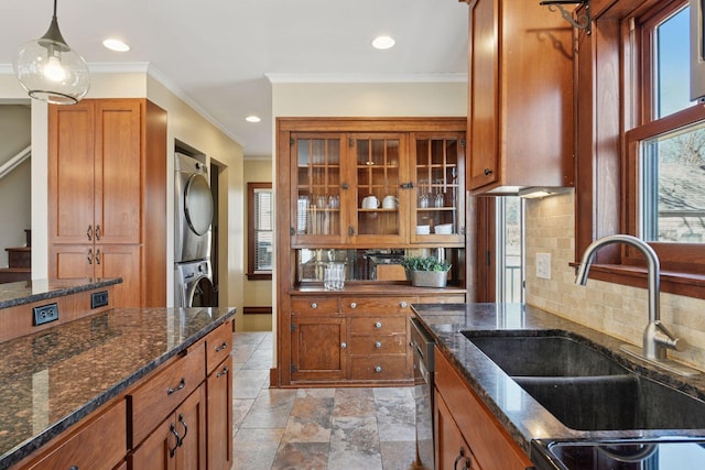 kitchen featuring brown cabinetry, dark stone counters, a sink, glass insert cabinets, and stacked washer / dryer