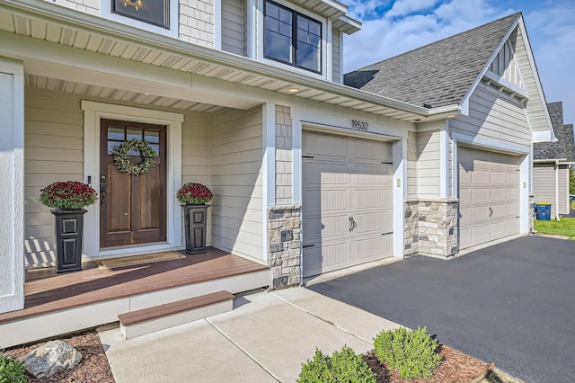 entrance to property featuring stone siding, driveway, an attached garage, and roof with shingles