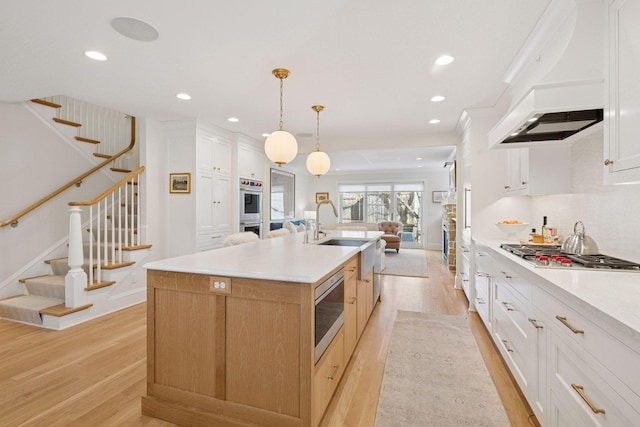 kitchen featuring custom range hood, tasteful backsplash, stainless steel appliances, light wood-style floors, and a large island with sink