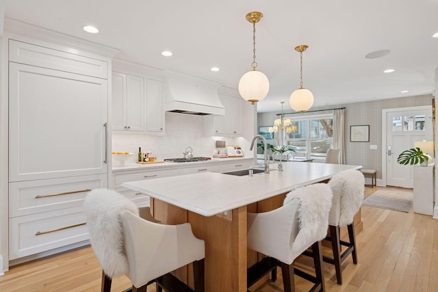 kitchen featuring a kitchen island with sink, a sink, light countertops, custom exhaust hood, and stainless steel gas cooktop