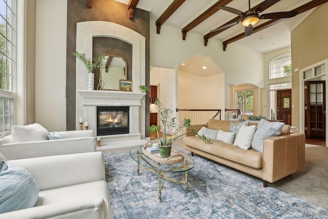 living area featuring beam ceiling, a glass covered fireplace, a high ceiling, and ceiling fan