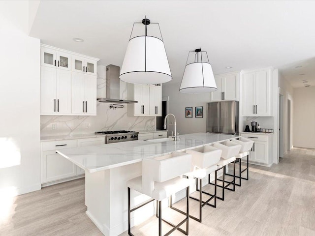 kitchen featuring backsplash, wall chimney range hood, freestanding refrigerator, light wood-style floors, and a kitchen island with sink