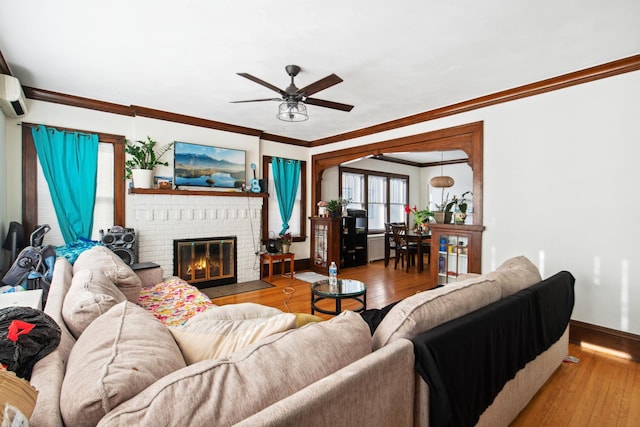 living room featuring a ceiling fan, wood-type flooring, crown molding, baseboards, and a brick fireplace