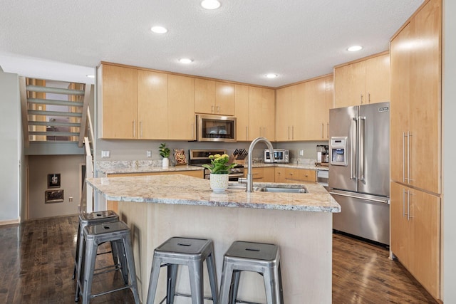 kitchen with a breakfast bar area, an island with sink, light brown cabinetry, stainless steel appliances, and dark wood-type flooring