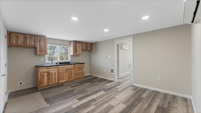 kitchen featuring a wall unit AC, brown cabinetry, wood finished floors, baseboards, and dark countertops