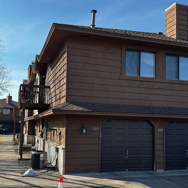 view of side of property with a garage, roof with shingles, central AC unit, and a balcony