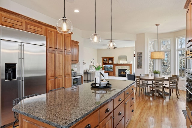 kitchen with a warm lit fireplace, brown cabinetry, light wood-type flooring, and appliances with stainless steel finishes