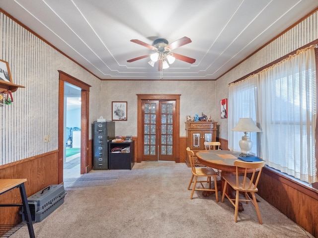 carpeted dining space featuring wallpapered walls, a wainscoted wall, ornamental molding, french doors, and a ceiling fan