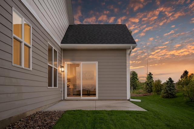 back of house at dusk featuring a patio area, a shingled roof, and a yard