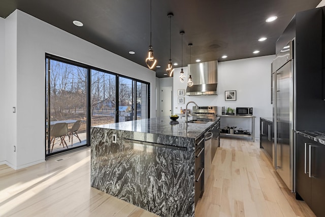 kitchen with an island with sink, dark stone countertops, light wood-style floors, wall chimney exhaust hood, and a sink