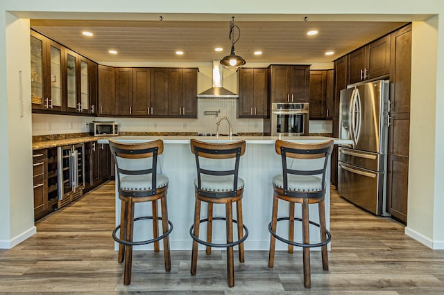 kitchen with a kitchen island with sink, wood finished floors, stainless steel appliances, dark brown cabinetry, and wall chimney exhaust hood