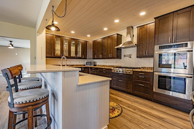 kitchen featuring light wood finished floors, wall chimney range hood, a kitchen breakfast bar, wooden ceiling, and stainless steel appliances