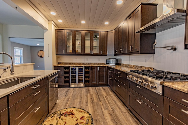 kitchen with a sink, stainless steel appliances, wine cooler, dark brown cabinets, and wall chimney exhaust hood