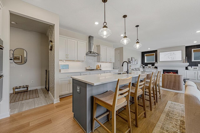 kitchen with light wood-style flooring, a warm lit fireplace, a sink, wall chimney range hood, and tasteful backsplash