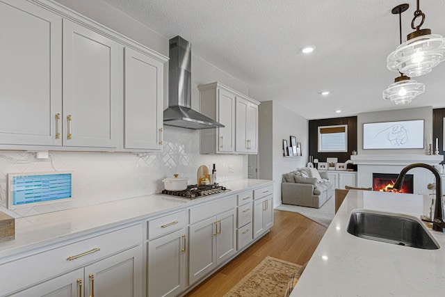 kitchen featuring wall chimney range hood, open floor plan, a warm lit fireplace, stainless steel gas stovetop, and a sink