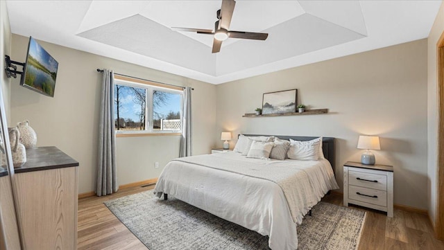 bedroom featuring a tray ceiling, light wood-style floors, baseboards, and ceiling fan