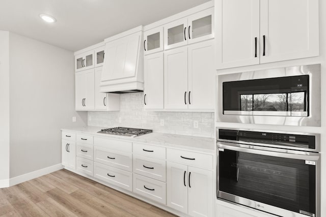 kitchen with backsplash, light stone countertops, light wood-type flooring, custom exhaust hood, and stainless steel appliances