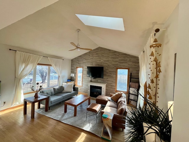 living room with ceiling fan, a skylight, wood finished floors, a glass covered fireplace, and high vaulted ceiling