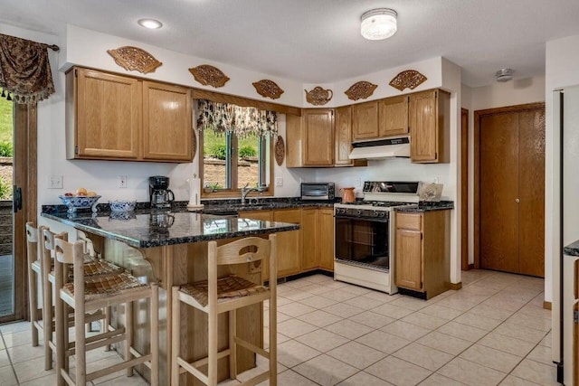 kitchen with range with gas stovetop, dark stone countertops, light tile patterned floors, under cabinet range hood, and a kitchen breakfast bar