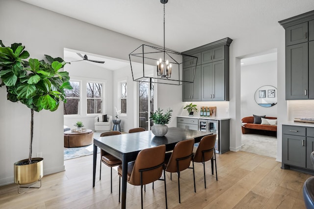 dining space featuring ceiling fan with notable chandelier, beverage cooler, baseboards, and light wood finished floors