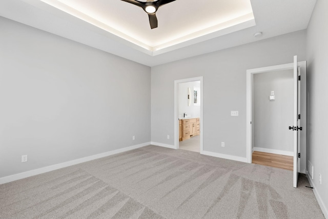 unfurnished bedroom featuring ceiling fan, baseboards, light carpet, ensuite bathroom, and a raised ceiling