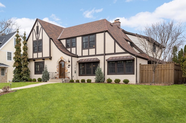 tudor-style house featuring a front lawn, stucco siding, a chimney, and fence