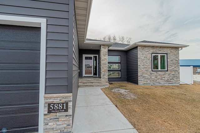 doorway to property with a garage, a lawn, stone siding, and roof with shingles