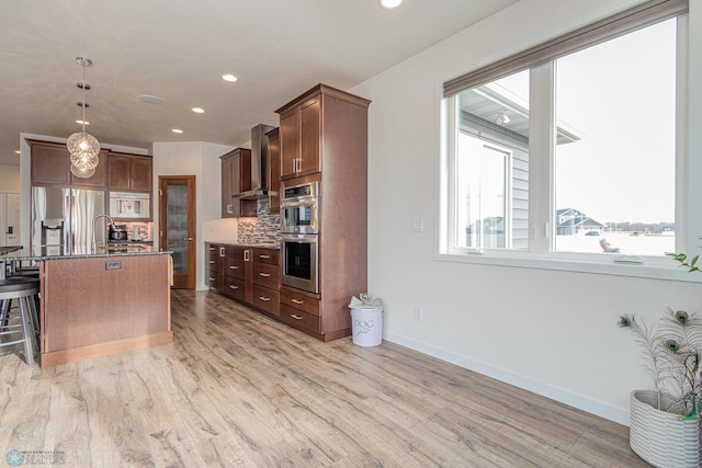 kitchen with light wood-style flooring, decorative backsplash, hanging light fixtures, appliances with stainless steel finishes, and wall chimney range hood