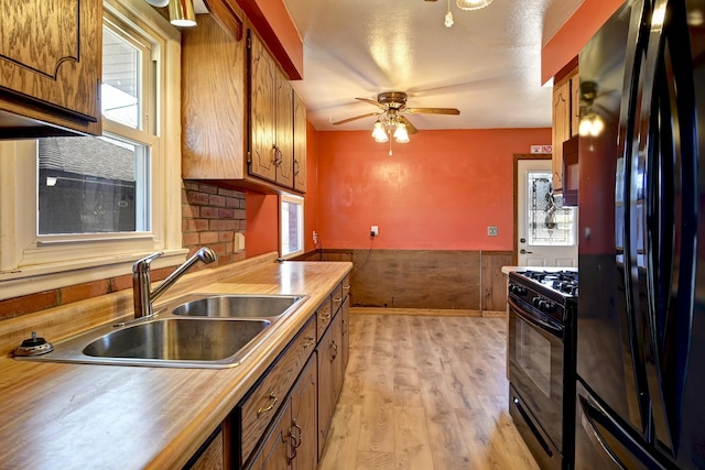 kitchen featuring light wood finished floors, wainscoting, brown cabinetry, black appliances, and a sink