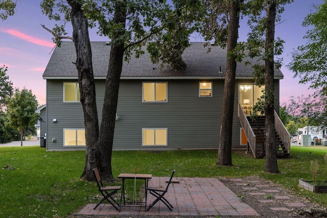 back of house at dusk featuring stairs, a patio, a yard, and roof with shingles