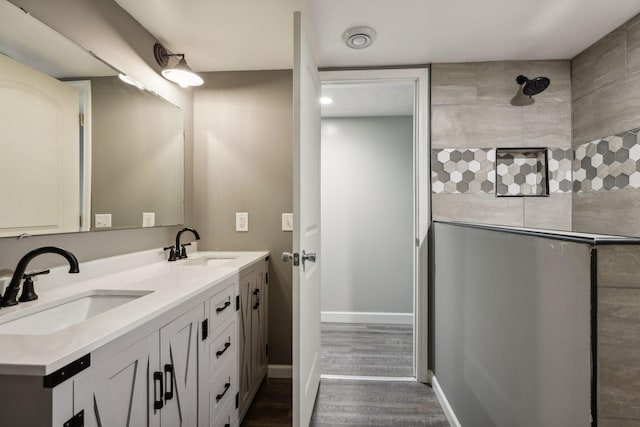 bathroom featuring baseboards, double vanity, a tile shower, wood finished floors, and a sink