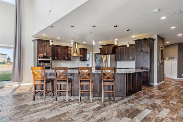 kitchen featuring a large island, recessed lighting, appliances with stainless steel finishes, wall chimney exhaust hood, and dark brown cabinets