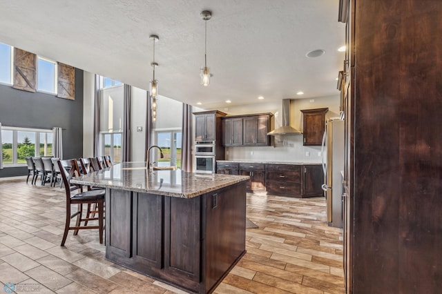 kitchen with a breakfast bar, an island with sink, stainless steel appliances, dark brown cabinets, and wall chimney exhaust hood