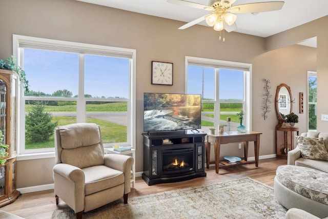 living area featuring a glass covered fireplace, a ceiling fan, baseboards, and wood finished floors