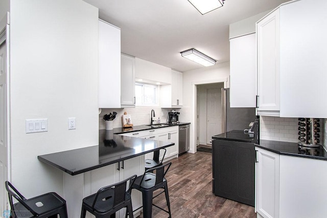 kitchen with a sink, a kitchen breakfast bar, stainless steel dishwasher, dark countertops, and white cabinetry