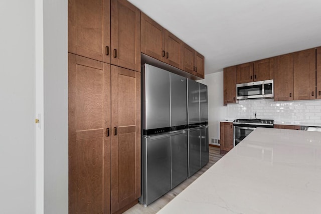 kitchen with light stone counters, brown cabinetry, stainless steel appliances, decorative backsplash, and light wood-type flooring