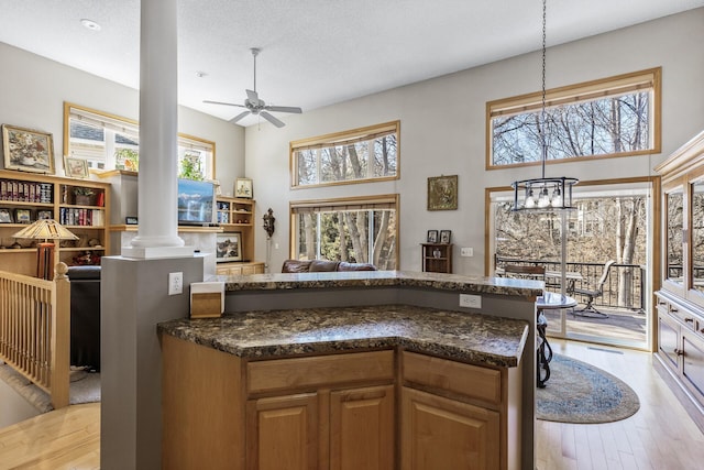 kitchen with dark stone countertops, ceiling fan with notable chandelier, light wood-style floors, and decorative columns