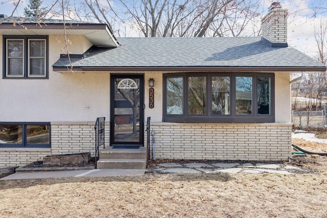 view of exterior entry with stucco siding, a chimney, brick siding, and a shingled roof