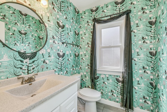 half bathroom featuring baseboards, toilet, vanity, and wallpapered walls