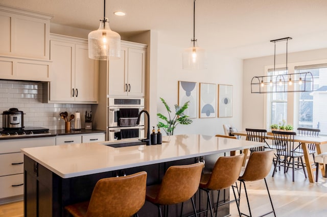 kitchen with backsplash, a center island with sink, double oven, stovetop, and a sink